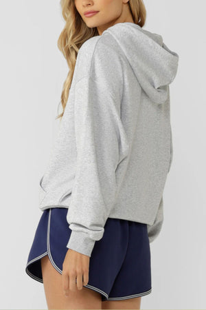 A person with long, wavy blonde hair wears a Lorna Jane Active Club Hoodie with a boucle applique logo and navy blue athletic shorts with white trim, standing against a plain light background.