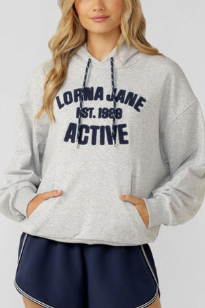 A woman wears a light gray Lorna Jane Active Club Hoodie with "LORNA JANE EST. 1989 ACTIVE" on the front, paired with navy blue athletic shorts featuring white trim.