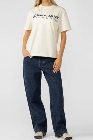 A person stands with one hand in their pocket against a plain background, wearing an oversized cream Lorna Jane Active Living T-Shirt, loose dark blue jeans, and black-and-white sneakers—an essential for active living.