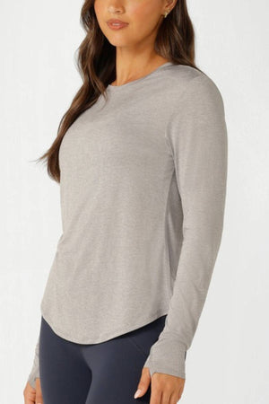 A woman wears the Lorna Jane Amy Dynamic Active Long Sleeve T-Shirt with dark leggings, standing against a white background and facing slightly to the side—ideal for lightweight layering.