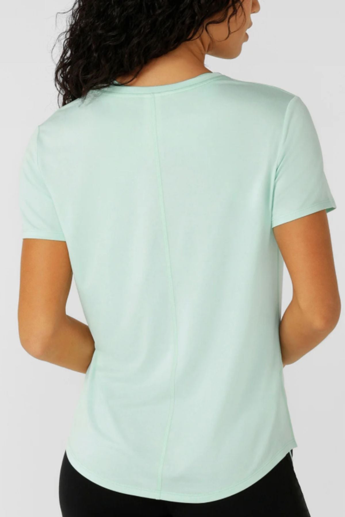 A woman with curly hair is seen from the back, wearing a light blue Lorna Jane Amy Dynamic Active T-Shirt and black pants against a plain light background. This quick-drying top is ideal for women’s workouts.