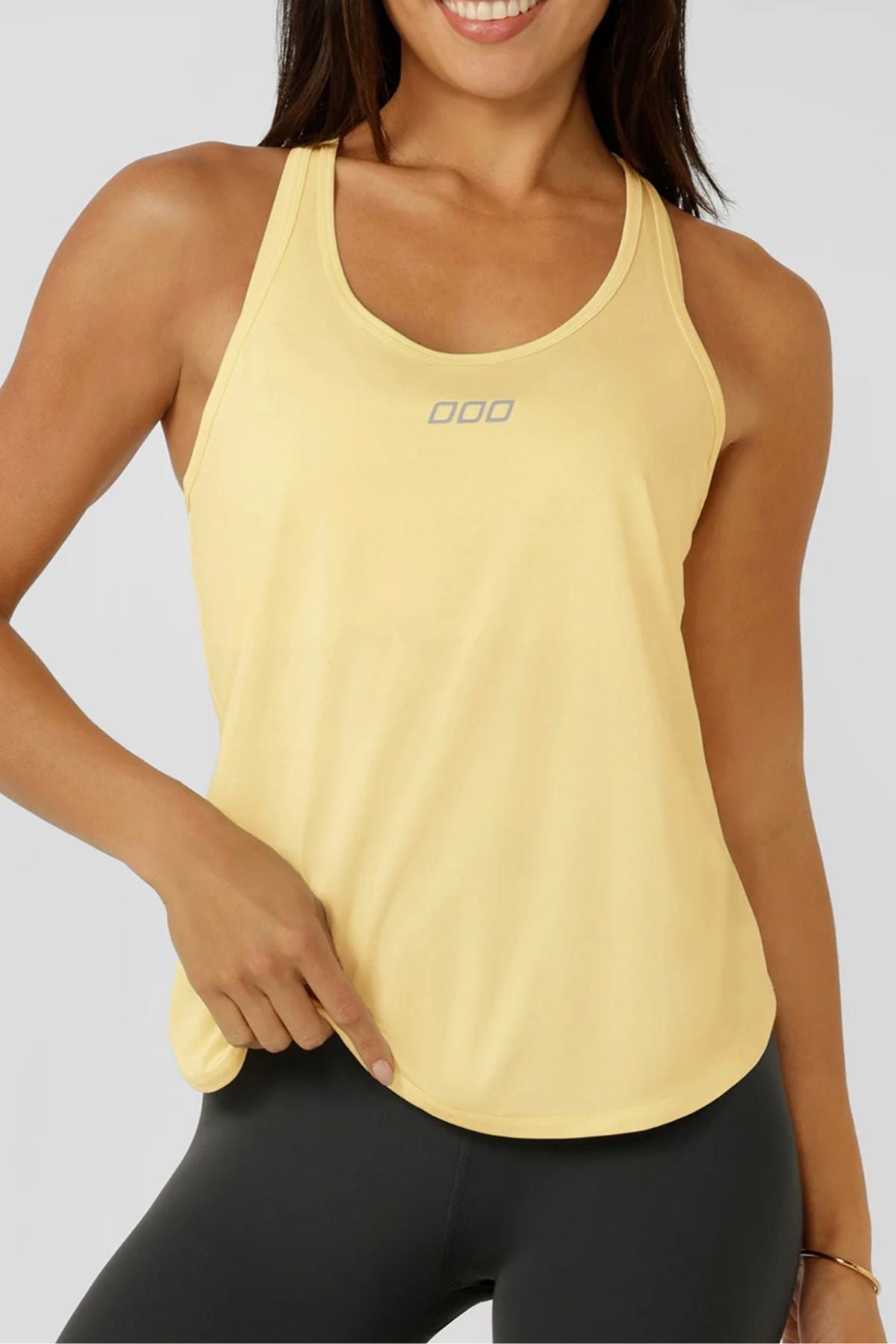 A smiling woman in black leggings points to the hem of her Lorna Jane Amy Dynamic Active Tank, which is light yellow with a small chest logo and made from quick-drying fabric for maximum comfort.