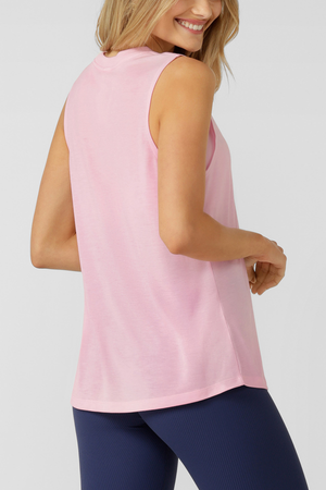 A smiling blonde woman poses sideways against a plain light background, wearing a Lorna Jane Lotus Muscle Tank in light pink paired with navy blue leggings.