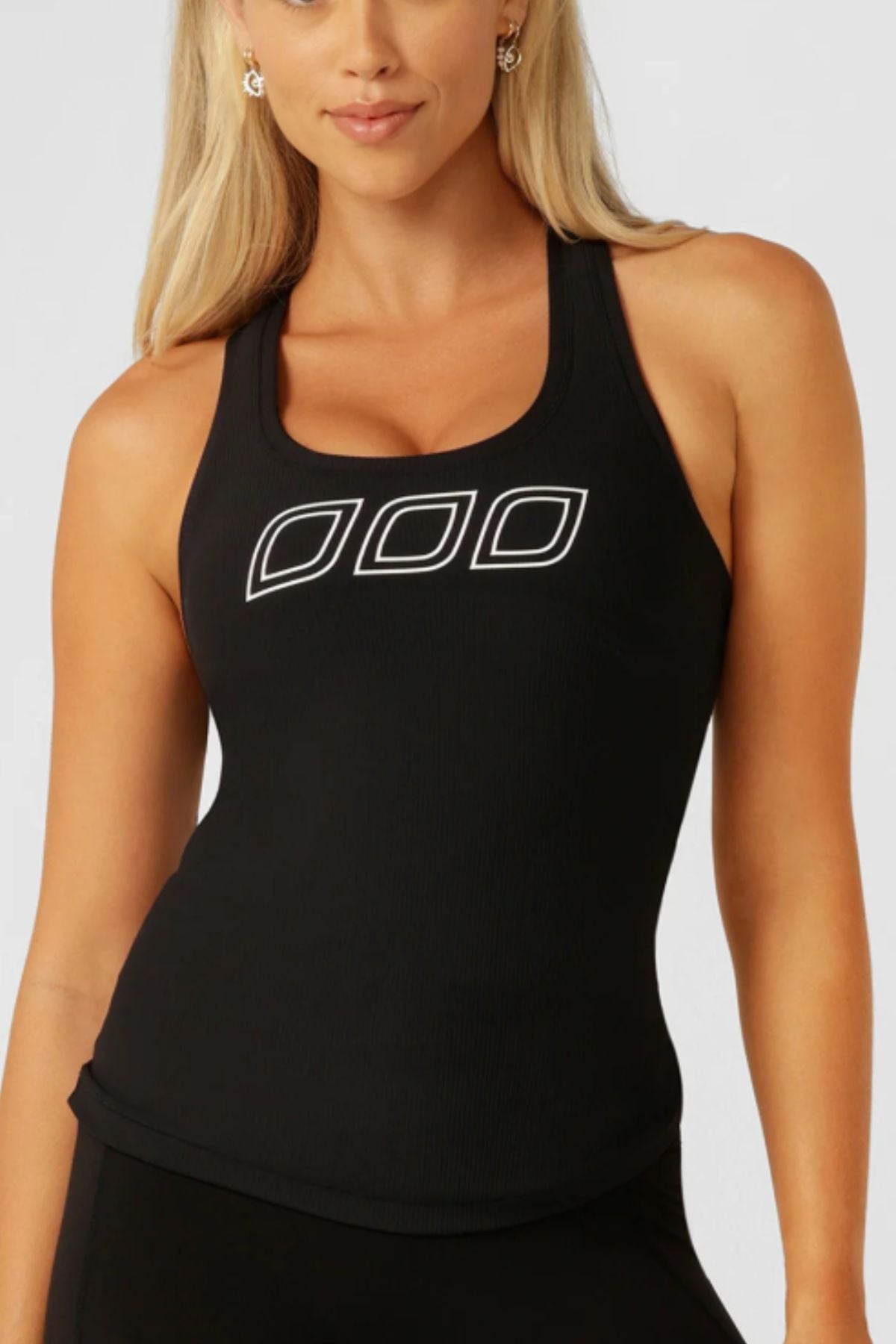 A woman with long blonde hair wears the Lorna Jane Amy Fitted Active Rib Tank, a black racerback style featuring three white-outlined geometric shapes on the chest. She stands facing forward against a plain light background.