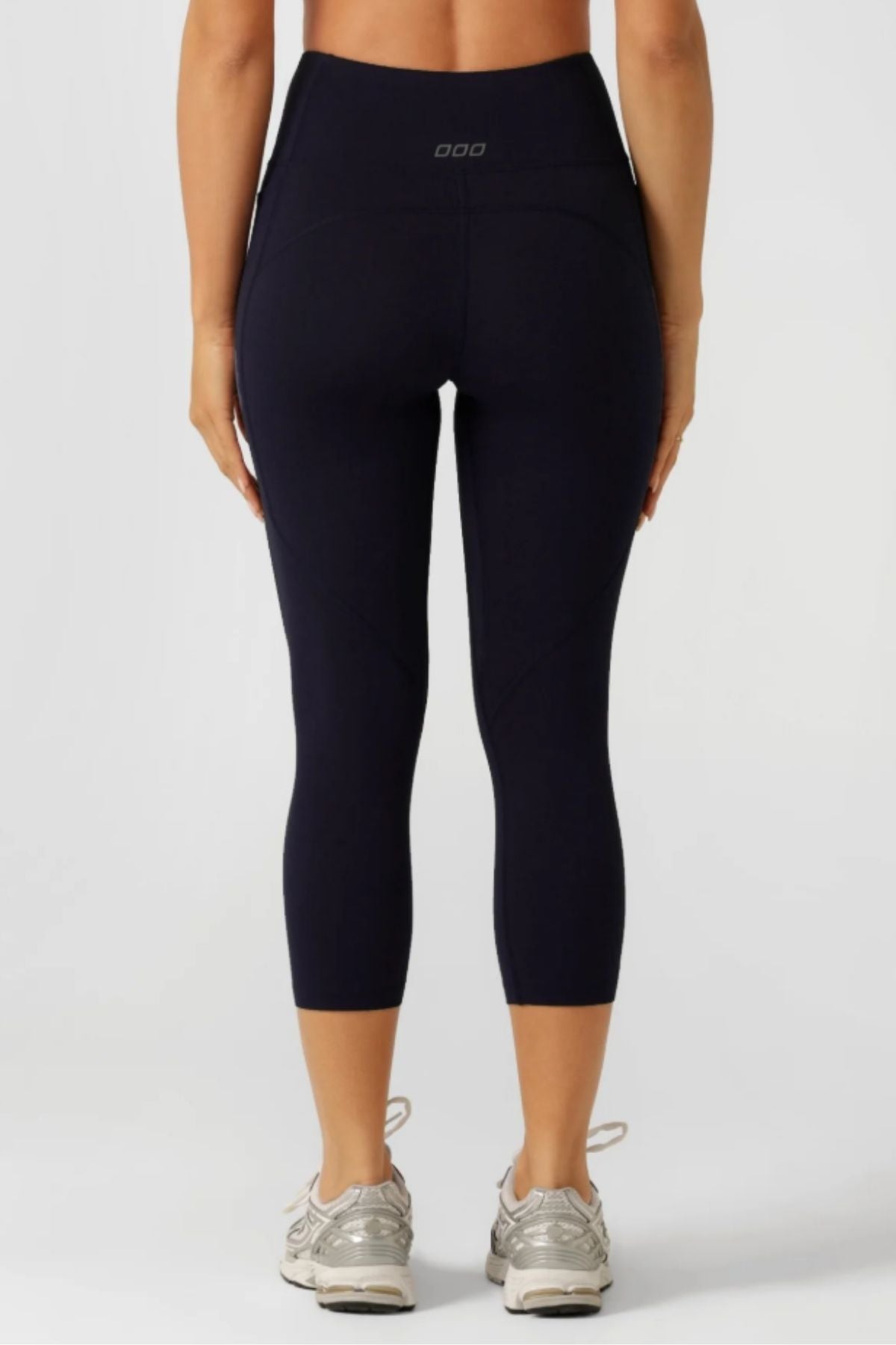 A woman is shown from the back wearing Lorna Jane Amy No Ride Phone Pocket 7/8 Leggings in black with Active Core Stability, paired with light sneakers, standing on a plain, light background.