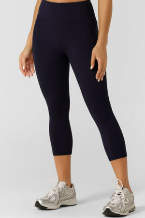 A person models the Lorna Jane Amy No Ride Phone Pocket 7/8 Leggings in black with moisture-wicking fabric and silver running shoes, standing against a plain light gray background. The photo is cropped to show only the lower half of the body.