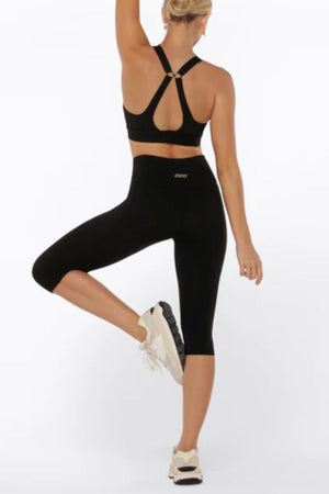A woman models the Lorna Jane Amy Phone Pocket 3/4 Tech Leggings and a black sports bra, standing on one leg with her arm raised. She faces away, wearing white sneakers, against a plain white background.