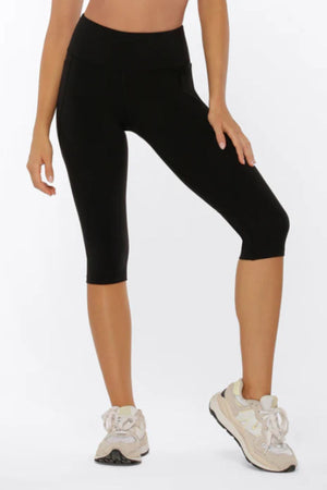 A person wearing black Lorna Jane Amy Phone Pocket 3/4 Tech Leggings and white sneakers stands with one knee slightly bent against a plain white background.