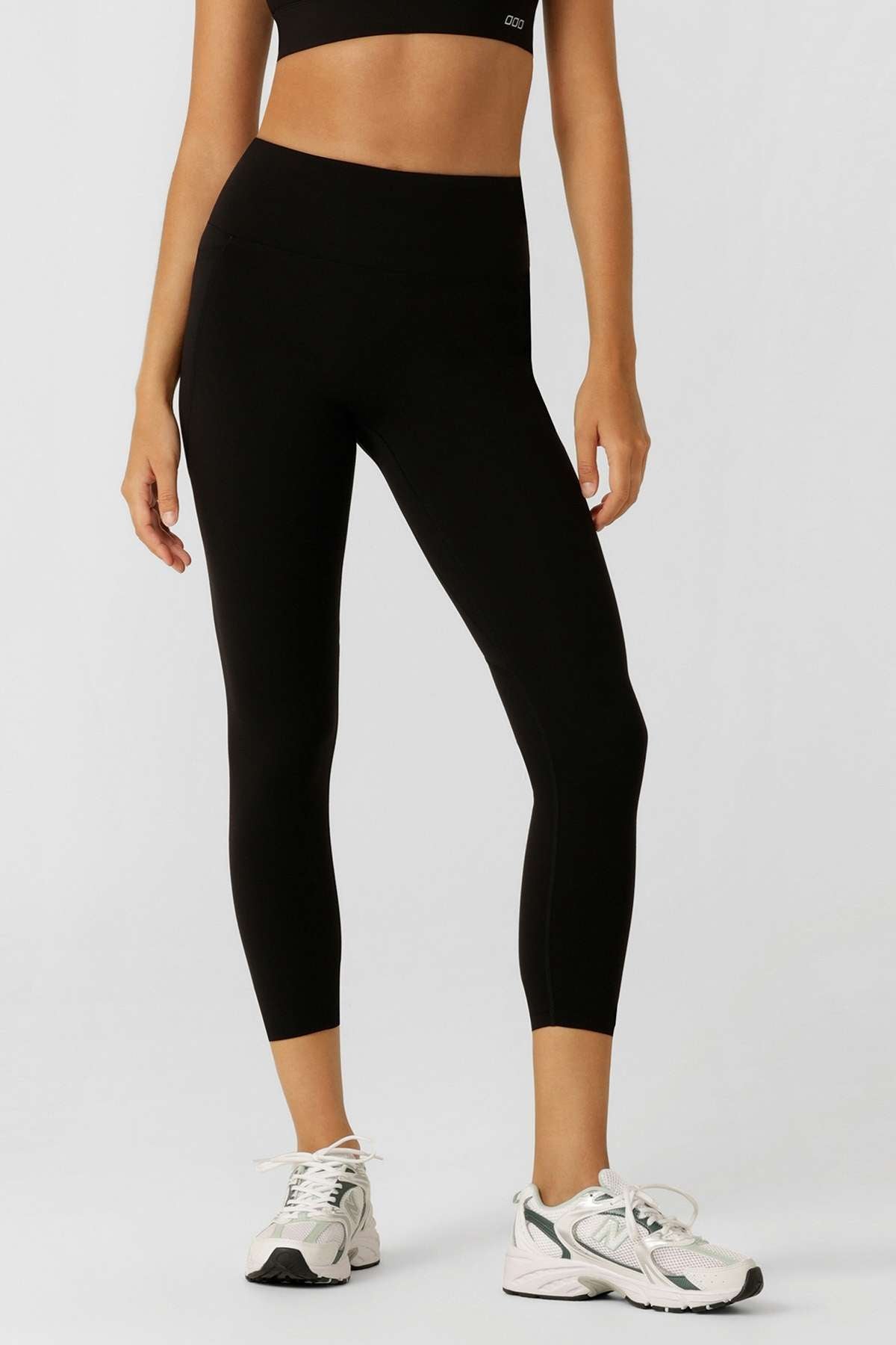 A person stands against a plain light background, arms relaxed at their sides, wearing a black sports bra, white athletic shoes, and Lorna Jane Amy Phone Pocket 7/8 Tech Leggings made with moisture-wicking fabric.