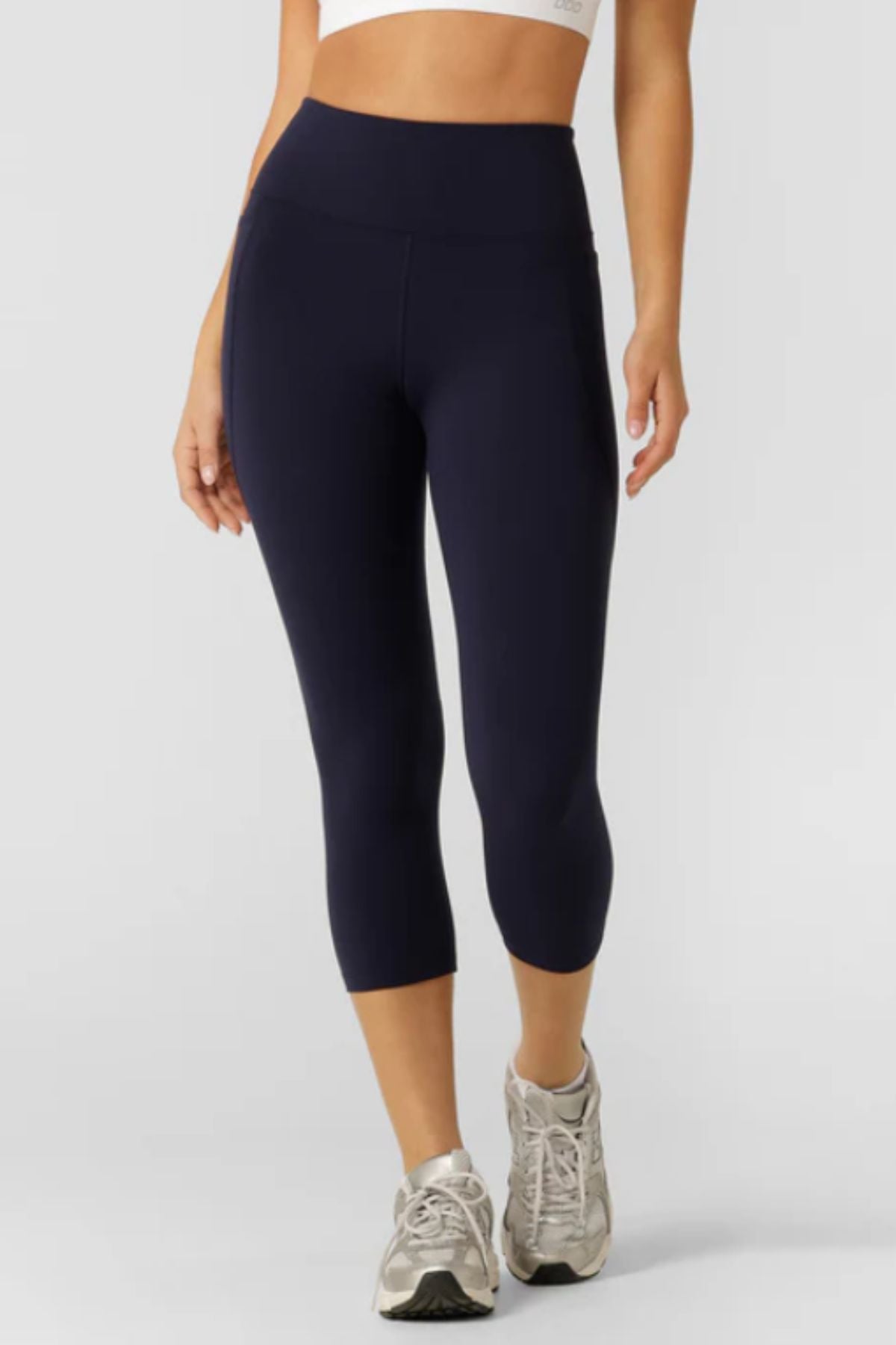 A person stands against a light background wearing navy Lorna Jane Amy Phone Pocket 7/8 Tech Leggings made from moisture-wicking fabric, paired with gray athletic shoes and a white sports bra peeking through.