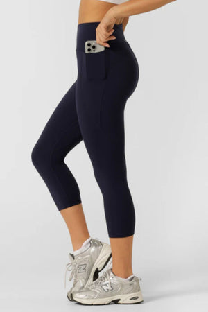 A person wearing navy blue Lorna Jane Amy Phone Pocket 7/8 Tech Leggings with a smartphone in the side pocket and silver athletic shoes, standing on a light background. These high-rise leggings feature moisture-wicking fabric for comfort.