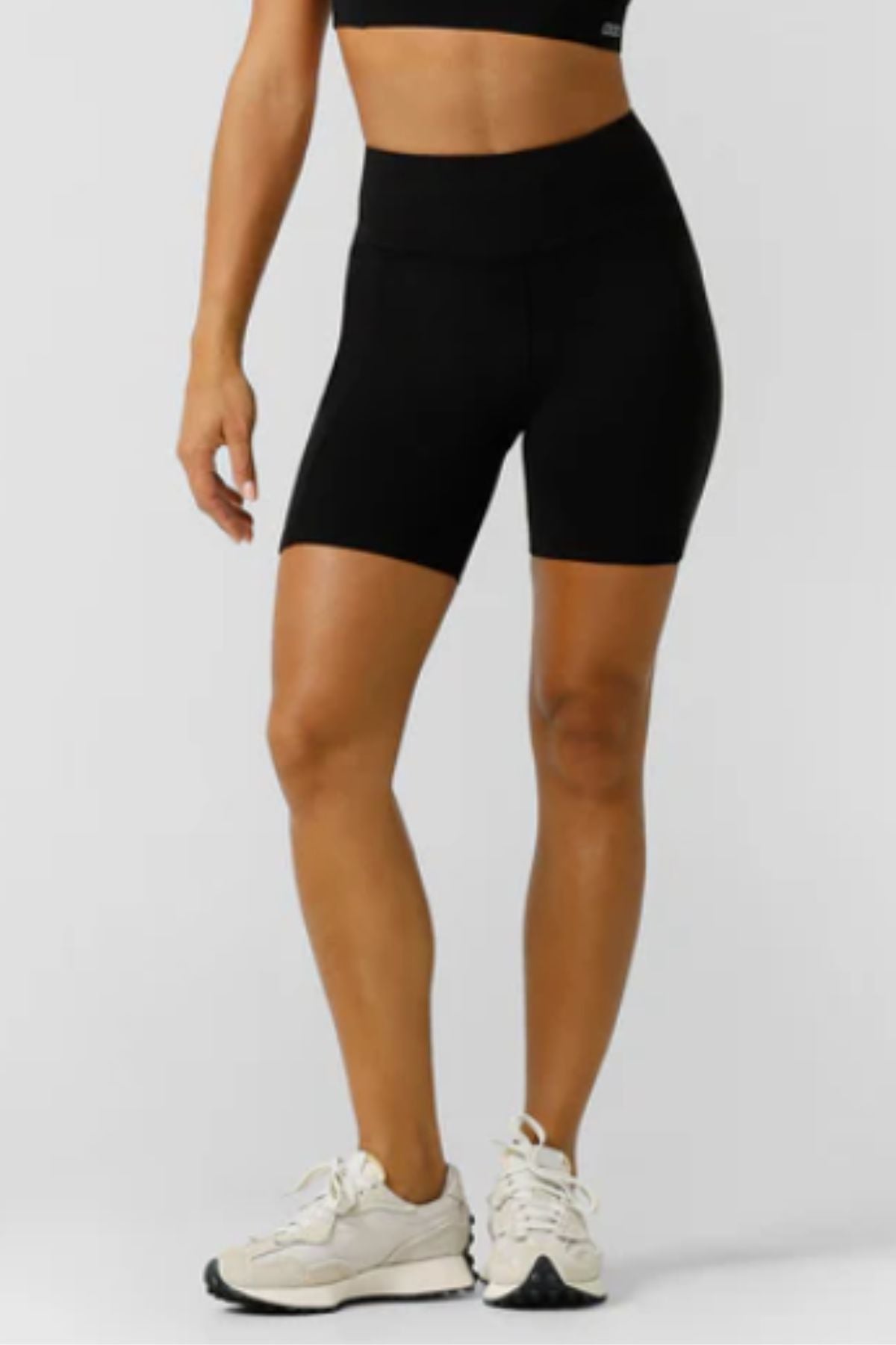 A person, visible from the shoulders down, wears a black sports bra, Lorna Jane Amy Phone Pocket Tech Bike Shorts with 4-way stretch, and white athletic shoes, standing against a plain light background.