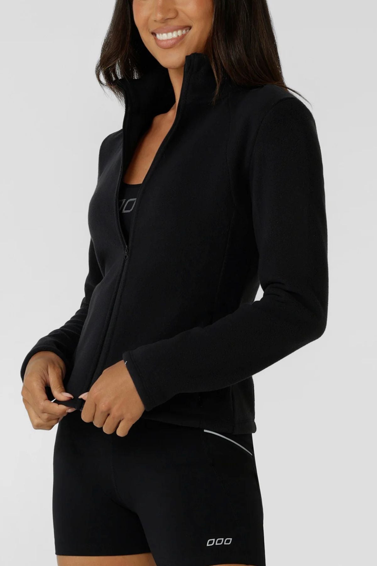 A woman smiles in a black Lorna Jane Amy Polar Fleece Zip Through Jacket over a black sports bra and shorts, with her hands near the zipper. The outfit has minimal white detail and is set against a plain light gray background.