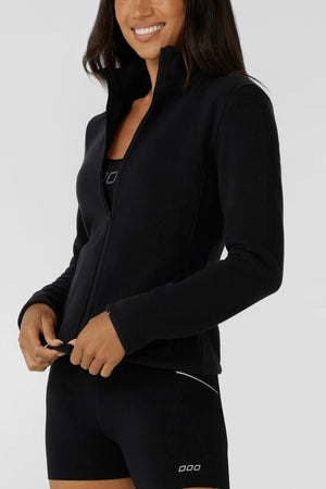 A woman smiles in a black Lorna Jane Amy Polar Fleece Zip Through Jacket over a black sports bra and shorts, with her hands near the zipper. The outfit has minimal white detail and is set against a plain light gray background.