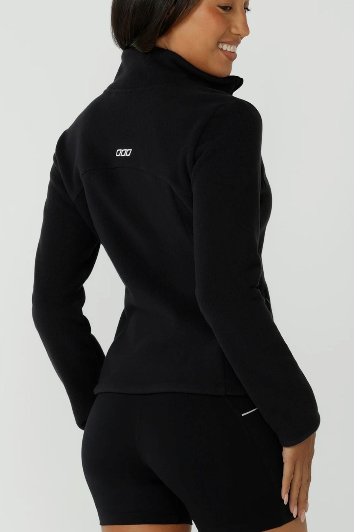 A woman wears a black Lorna Jane Amy Polar Fleece Zip Through Jacket and black shorts, standing with her back to the camera, smiling over her shoulder. The jacket features a small white logo on the back.