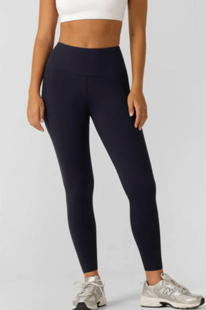 A person wearing a white sports bra, Lorna Jane Amy Thermal Phone Pocket Full Length Leggings for cold-weather workouts, and gray New Balance sneakers stands against a plain light gray background.