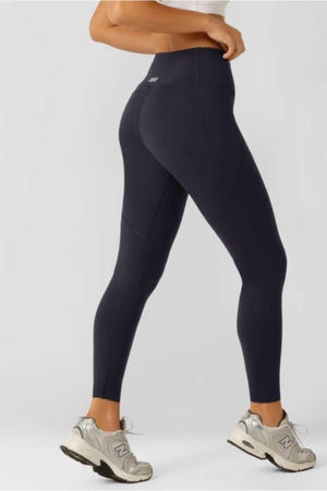 A person stands against a light background, showing only their lower half. They wear a white top, Lorna Jane Amy Thermal Phone Pocket Full Length Leggings in dark color for Active Core Stability, and light gray New Balance sneakers.