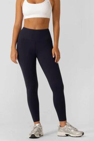 Wearing a white sports bra and Lorna Jane Amy Thermal Phone Pocket Full Length Leggings, paired with gray New Balance sneakers, a person stands against a plain light gray background.