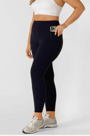 A person stands against a plain, light background wearing a white sports bra, silver athletic shoes, and Lorna Jane Amy Thermal Phone Pocket Full Length Leggings—perfect for cold-weather workouts.