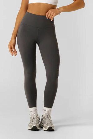 A person in a white crop top, Lorna Jane Amy Thermal Phone Pocket Full Length Leggings, white socks, and gray athletic shoes stands against a plain light background with one arm bent and the other relaxed.