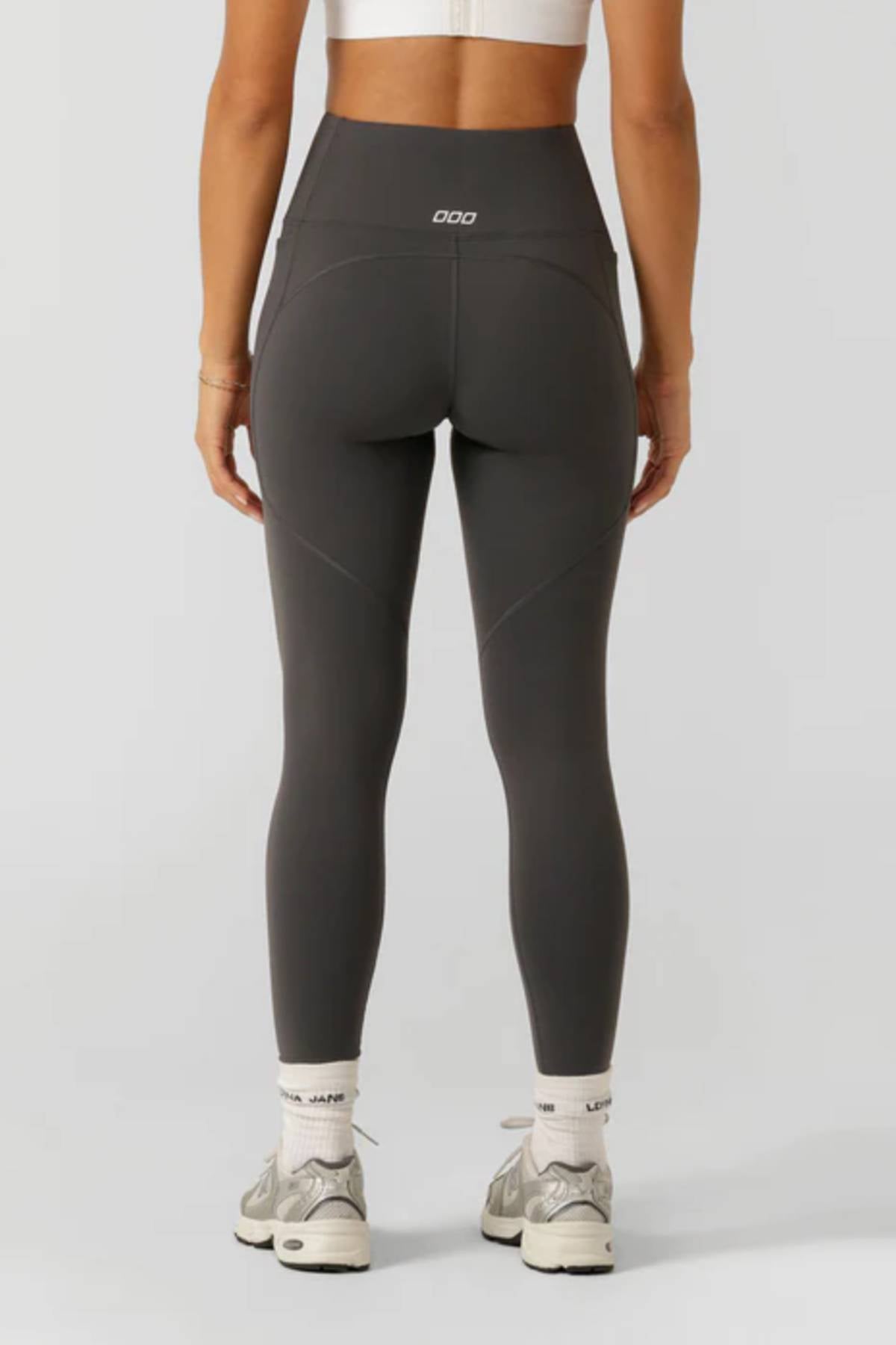A person in a white sports bra, Lorna Jane Amy Thermal Phone Pocket Full Length Leggings in dark gray, white socks, and gray sneakers stands with their back to the camera—ideal for cold-weather workouts.