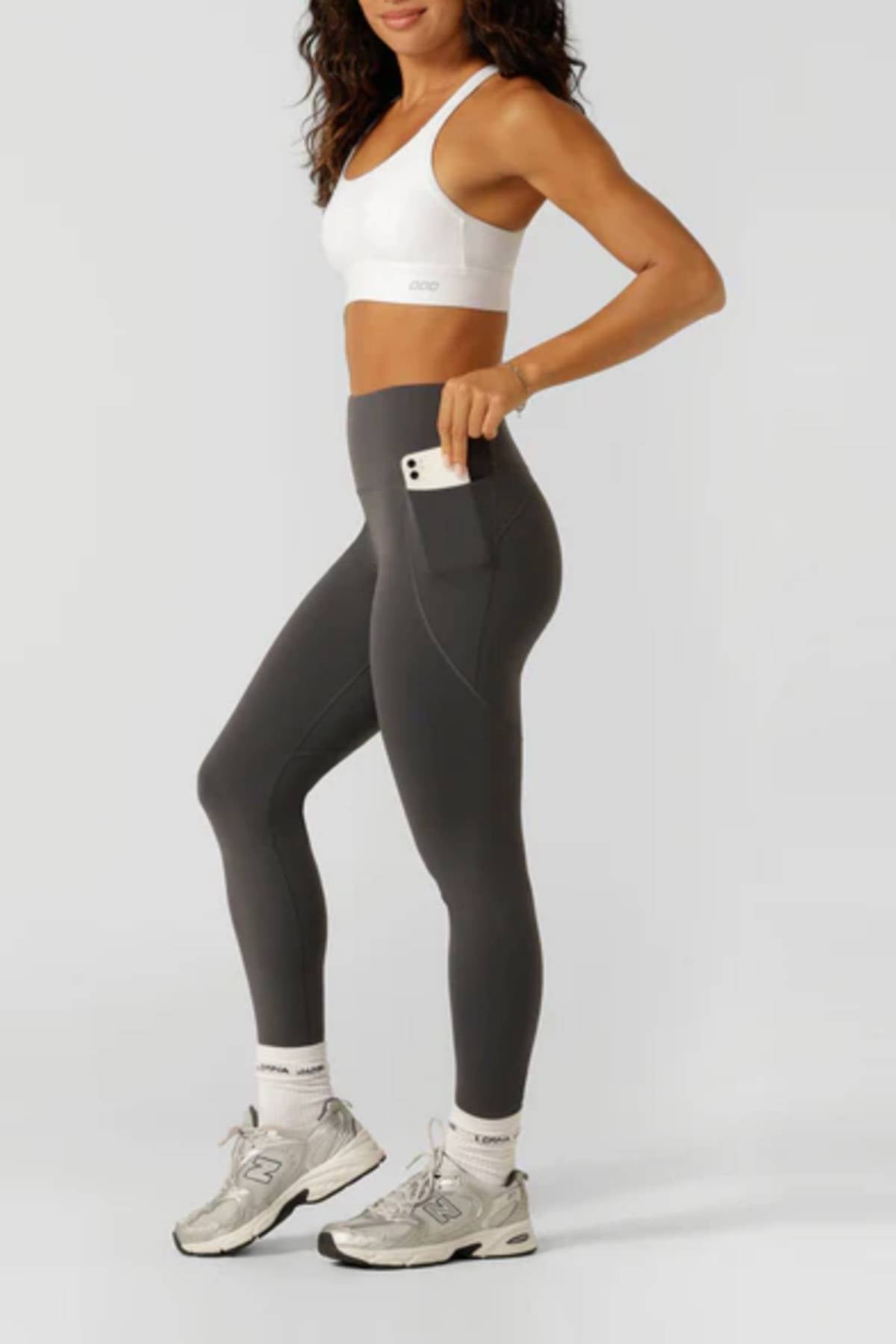 A woman in Lorna Jane Amy Thermal Phone Pocket Full Length Leggings and a white sports bra stands sideways, slipping her smartphone into the legging’s phone pocket—ideal for cold-weather workouts. She wears white socks and silver sneakers against a plain background.