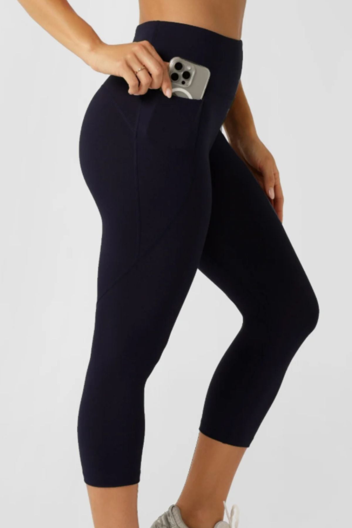 A woman wears Lorna Jane Amy No Ride Phone Pocket 7/8 Leggings, sliding her smartphone into the side pocket. Featuring Active Core Stability, she pairs them with light sneakers and stands against a plain background.