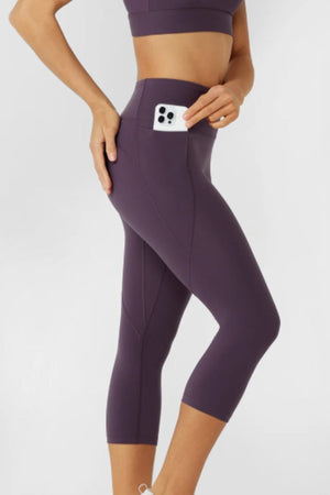 Wearing Lorna Jane Amy No Ride Phone Pocket 7/8 Leggings, a person stands against a plain background, matching a purple sports bra and tucking a phone into the leggings' waistband pocket.
