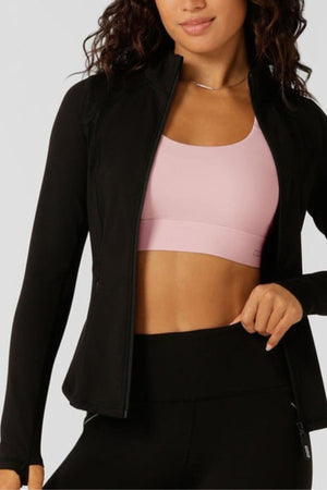 Woman wears the Lorna Jane Amy Thermal Active Zip Through Jacket with thumbholes over a light pink sports bra and black high-waisted leggings, standing with one hand holding her jacket. Her hair is wavy and she smiles slightly.
