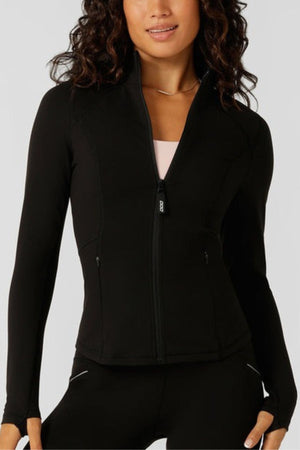 A woman wears the Lorna Jane Amy Thermal Active Zip Through Jacket—fitted, black, with sleek thumbholes—over a light pink top and matching black leggings. She has wavy dark hair and a slight smile.