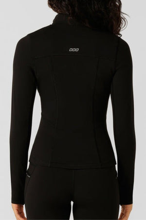 A woman with long dark hair wears the Lorna Jane Amy Thermal Active Zip Through Jacket and matching pants, viewed from behind. The black fitted jacket features a small white "ODD" logo on the upper back for a sleek, active look.