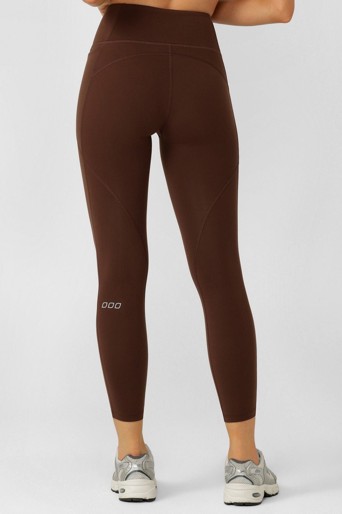 A person is shown from the back wearing Lorna Jane Amy No Chafe Phone Pocket Ankle Biter Leggings in brown with sweat-wicking fabric and gray sneakers, standing against a plain light-colored background.