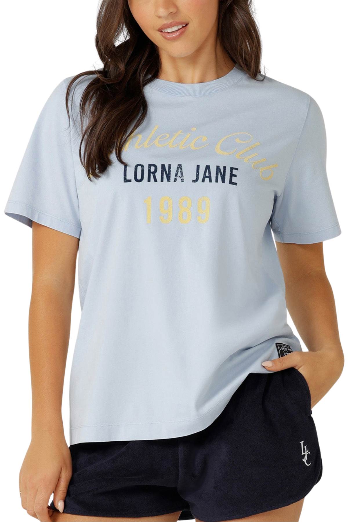 A woman stands with one hand in her pocket, wearing a relaxed-fit Lorna Jane Athletic Club Relaxed T-Shirt in light blue and dark blue shorts.