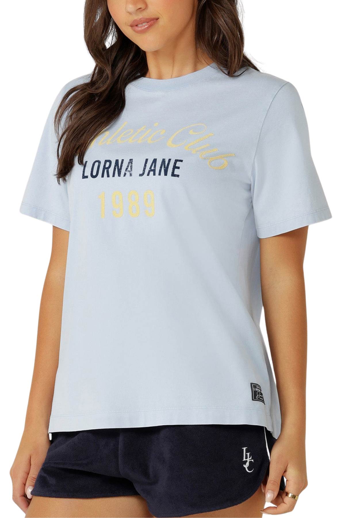 A woman wearing the Lorna Jane Athletic Club Relaxed T-Shirt in light blue and navy shorts with "LJ" initials stands against a white background, showcasing this vintage-inspired style in a classic relaxed fit.
