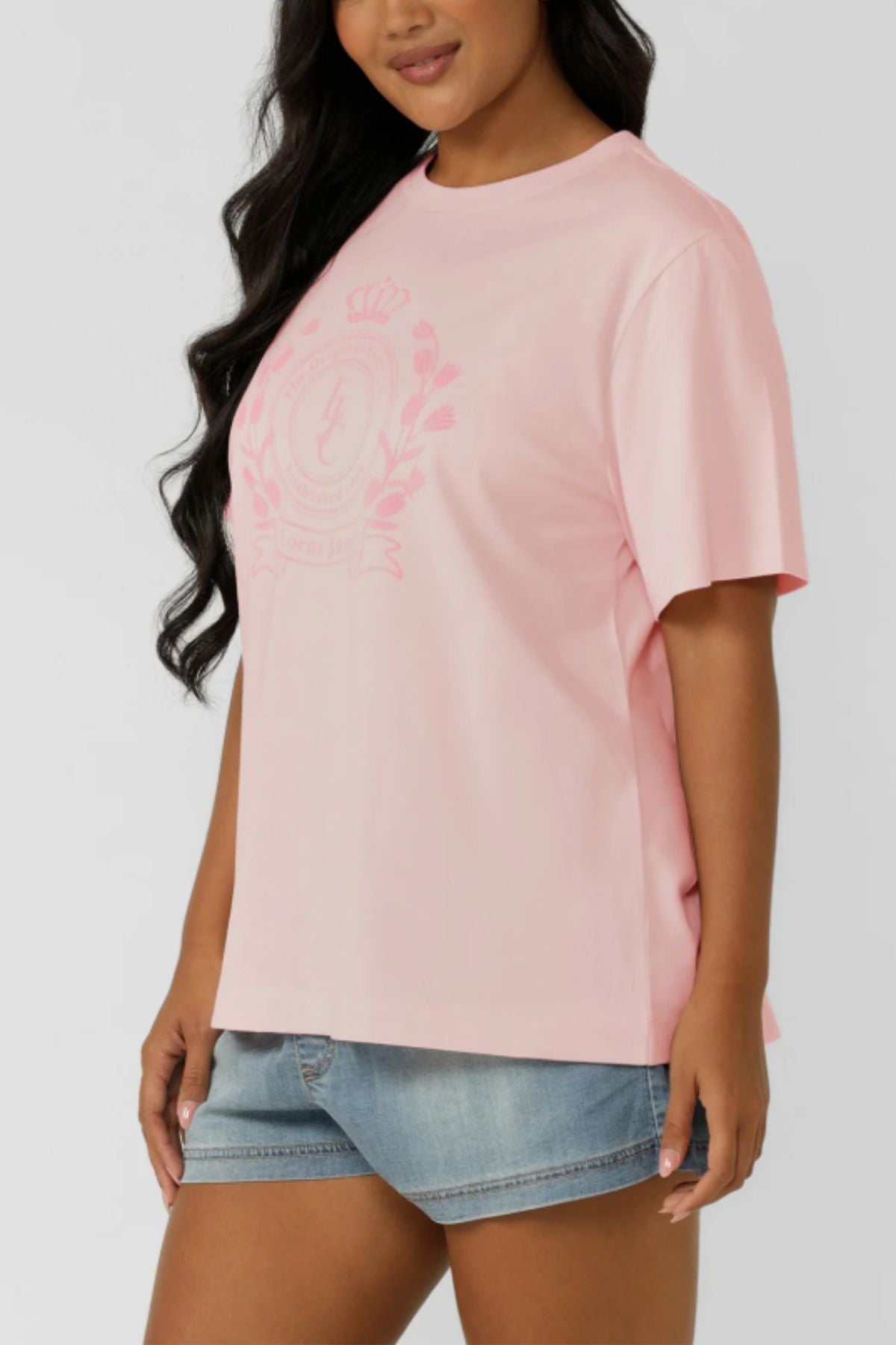 A woman in the Lorna Jane Chalet Relaxed T-Shirt—premium cotton, light pink, with a subtle front crest—paired with light blue denim shorts, stands against a plain background, embodying an active lifestyle.