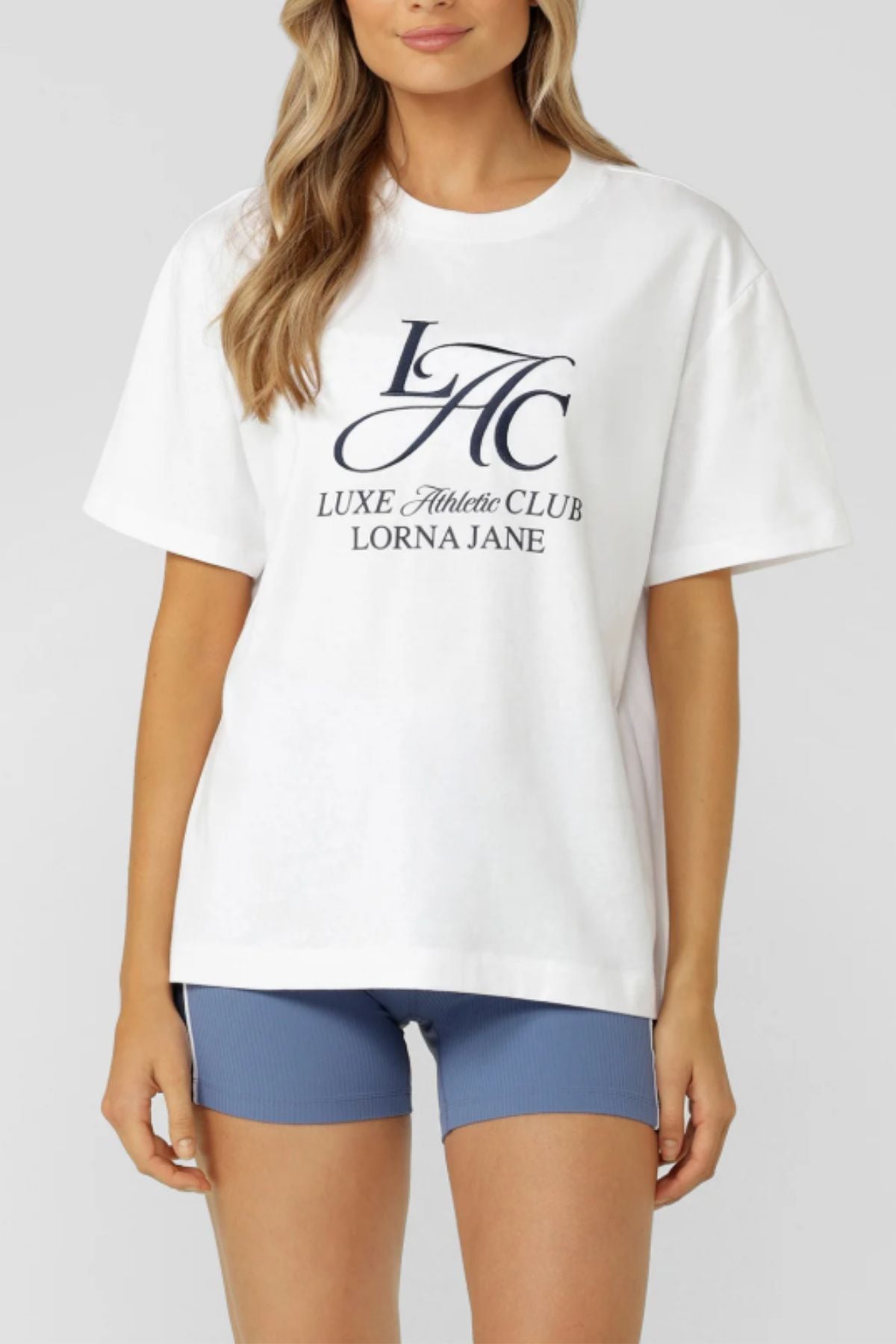 The woman wears the Lorna Jane Beaumont T-Shirt—an oversized, breathable cotton tee—paired with fitted blue athletic shorts, standing against a plain background.