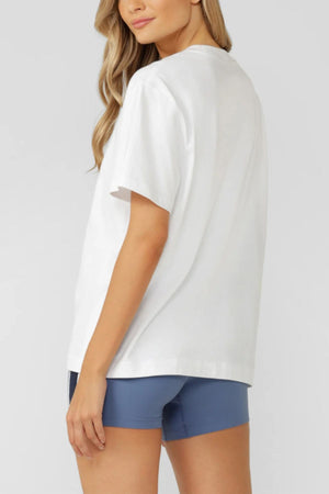 A woman with long, wavy blonde hair stands sideways, looking over her shoulder. She wears a Lorna Jane Beaumont T-Shirt and fitted blue shorts against a plain, light gray background.