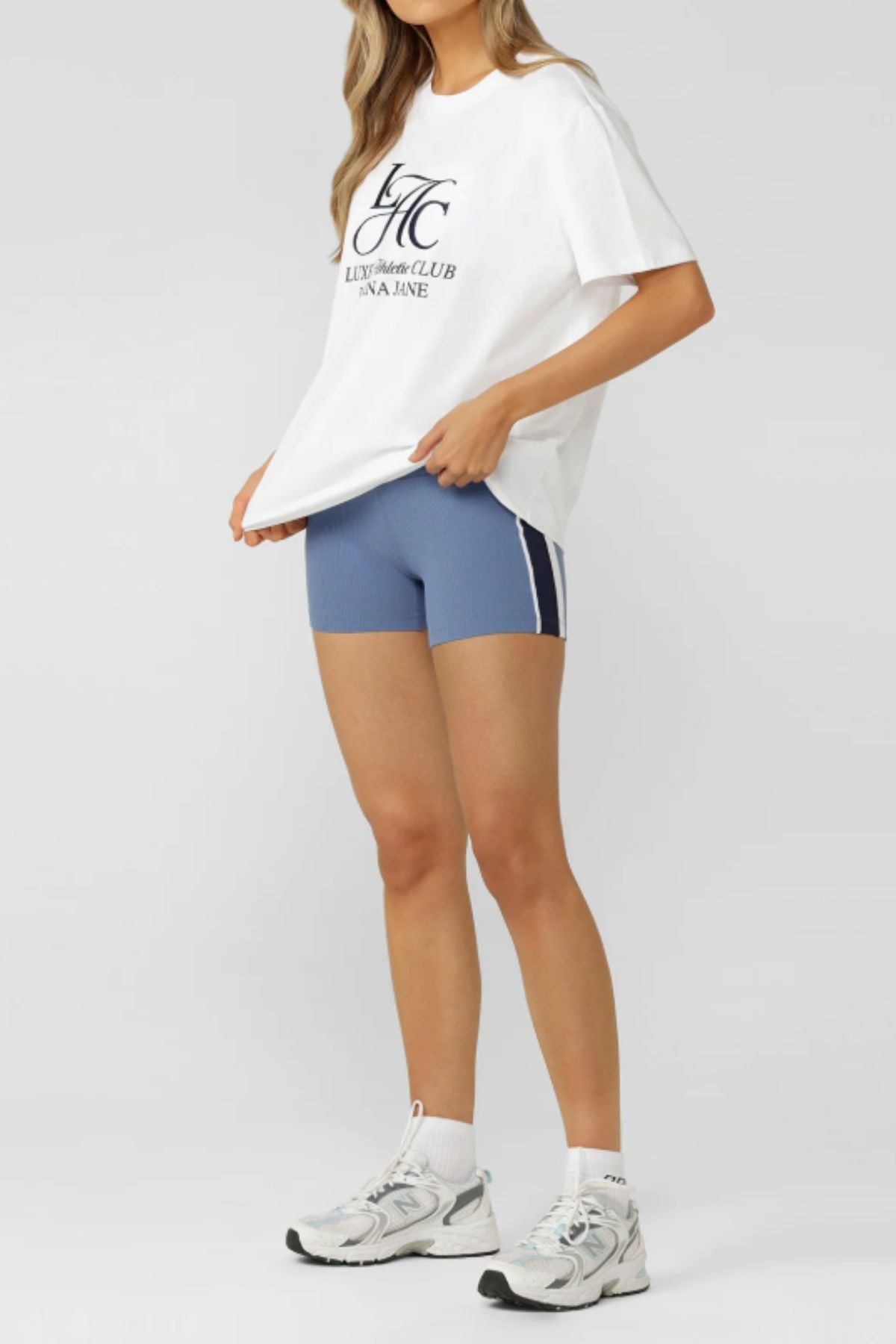 A person stands against a light gray background wearing a white Lorna Jane Beaumont T-Shirt made from breathable cotton, blue athletic shorts with white stripes, and white sneakers. Their face is not visible.