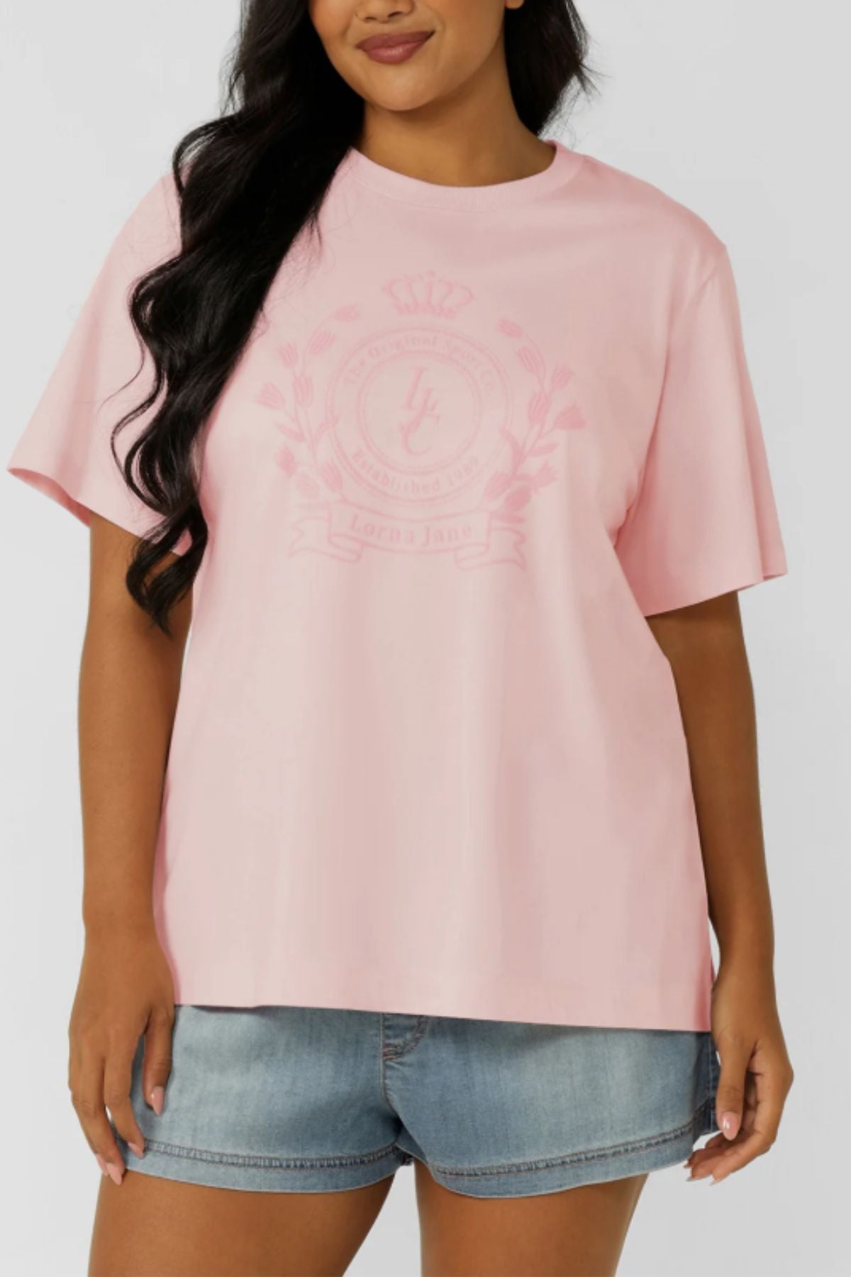 A woman stands against a plain background wearing the Lorna Jane Chalet Relaxed T-Shirt in light pink with a subtle emblem on premium cotton, paired with light denim shorts.