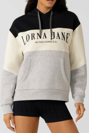 A woman wears the Lorna Jane Championship Oversized Fleece Hoodie, featuring “Active Living Co” on the front, bold black, cream, and gray color blocking, a drawstring hood, and front pocket. She pairs it with black shorts.