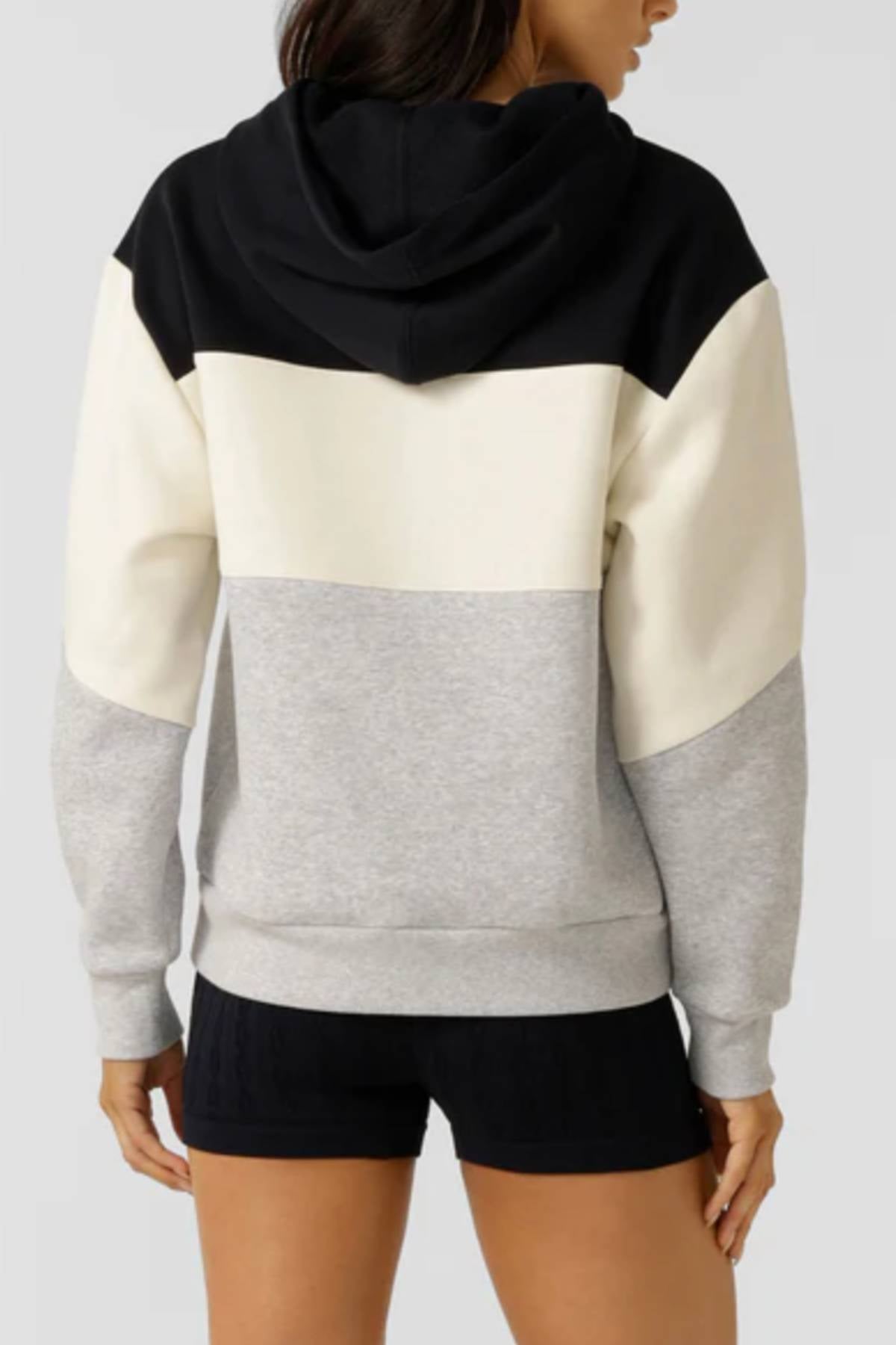 A woman facing away wears the Lorna Jane Championship Oversized Fleece Hoodie, featuring bold black, white, and gray color blocking, a hood, long sleeves, and paired with black shorts for a cozy look.