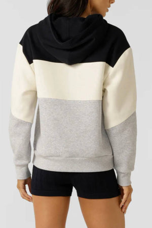 A woman facing away wears the Lorna Jane Championship Oversized Fleece Hoodie, featuring bold black, white, and gray color blocking, a hood, long sleeves, and paired with black shorts for a cozy look.