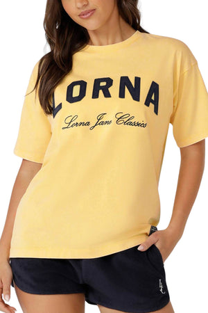 A woman wears an oversized yellow Lorna Jane Classics Washed Vintage T-Shirt with a statement front logo, paired with dark shorts. Her right hand rests in her shorts pocket.