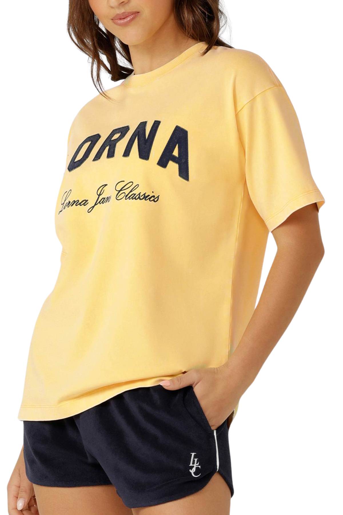 A woman wears the Lorna Jane Classics Washed Vintage T-Shirt, oversized in yellow with "LORNA" and "Lorna Jane Classics" logos, paired with black shorts featuring a small white logo. She rests her hand casually in her pocket.