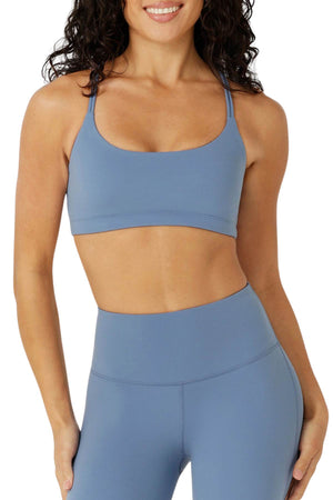 A woman models the Lorna Jane Lotus Cropped Sports Bra in light blue, paired with matching high-waisted leggings, smiling with her hand at her side. The photo is cropped from shoulders to upper thighs on a white background.
