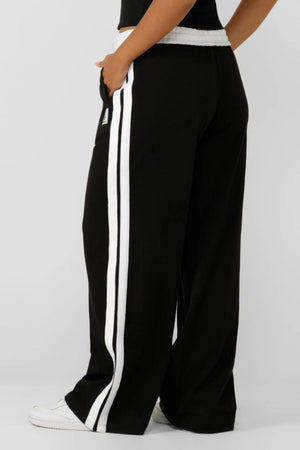 A person wears Lorna Jane Dance Pant—black wide-leg pants with white side stripes and a relaxed fit—paired with white sneakers, standing with one hand in their pocket against a plain background.