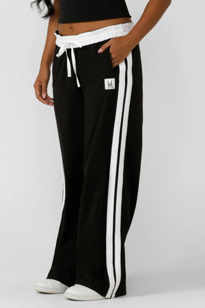 Someone is wearing the Lorna Jane Dance Pant—relaxed cut, black with white side stripes, a white adjustable drawstring waistband, and a small patch on the front pocket—paired with a black top and white sneakers.