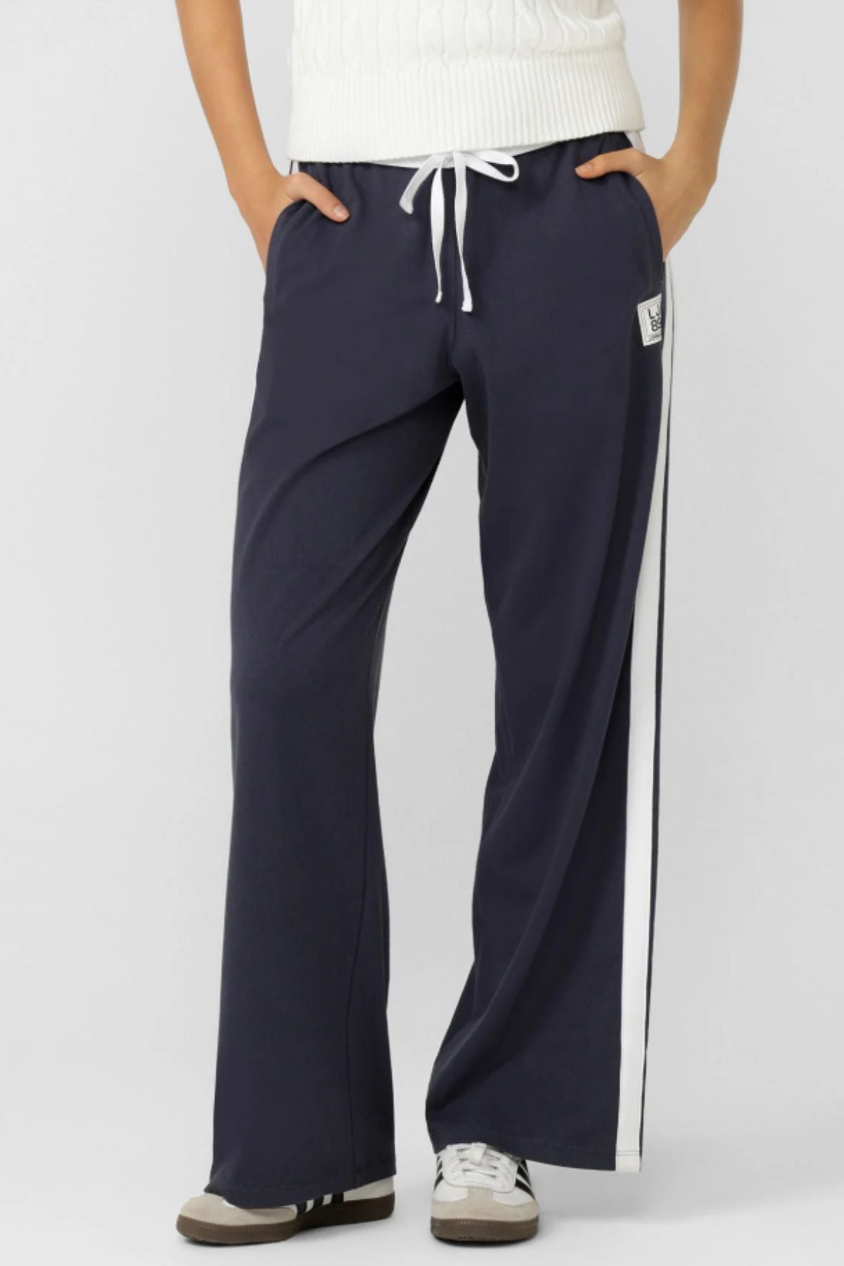 Wearing Lorna Jane Dance Pant sweatpants in retro sport navy blue with a white side stripe and adjustable waistband, paired with white sneakers and a white sweater, this person stands casually with hands in pockets.