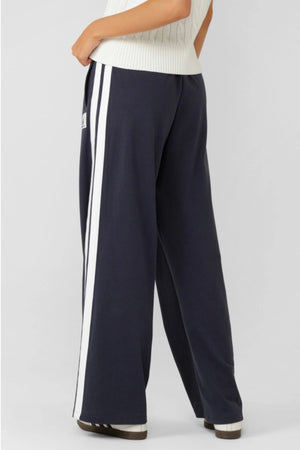 A person wearing Lorna Jane Dance Pant in retro sport navy blue with white side stripes and an adjustable waistband, paired with a white knit top, stands with one hand in their pocket, facing away from the camera.