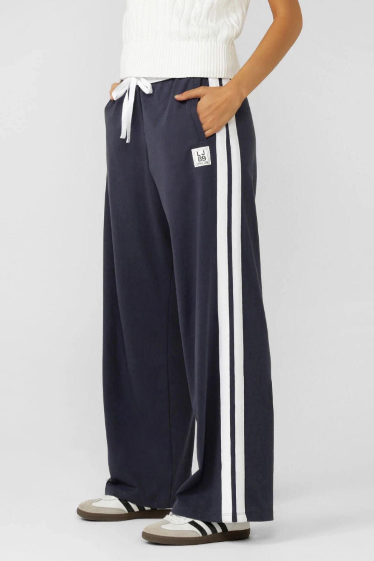 A person wears Lorna Jane Dance Pants in navy blue with a white adjustable waistband and side stripes for a retro sport vibe, paired with a white knit top and black-and-white sneakers.